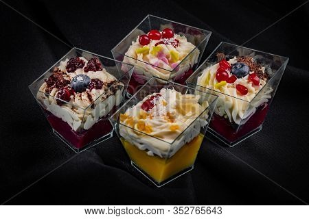 Four Trapezoid Glasses With Mixed Fruit-and-cream Desserts Of Different Flavours On Dark Background