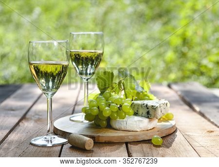 Glasses Of White Wine Near Delicious Cheese And Green Grape On Wooden Table With Green Plants On Blu