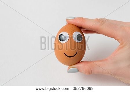Female Hand Holding Egg-smiley With Googly Eyes On White Background. Food With Funny Face.