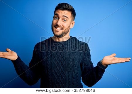 Young handsome man wearing casual sweater standing over isolated blue background smiling showing both hands open palms, presenting and advertising comparison and balance