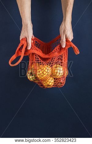 Apples And Lemons In An Orange String Bag In Men's Hands On A Dark Blue Background. Side View.