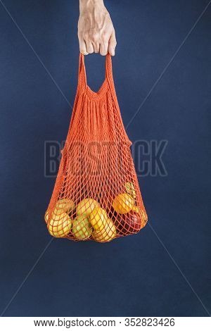 Apples And Lemons In An Orange String Bag In A Man's Hand On A Dark Blue Background. Side View.