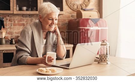 Happy Aged Woman Relaxing In Kitchen With Laptop And Coffee, Watchig Videos Or Browsing Internet, Pa