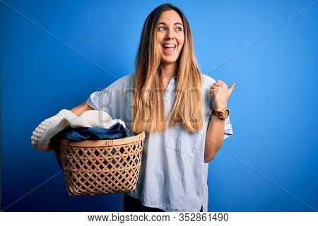 Young beautiful blonde woman doing housework holding wicker basket with clothes pointing and showing with thumb up to the side with happy face smiling