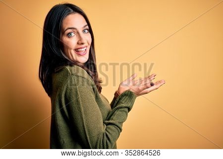 Young brunette woman with blue eyes wearing green casual sweater over yellow background pointing aside with hands open palms showing copy space, presenting advertisement smiling excited happy