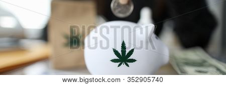 Close-up Of Person Hand Putting Coin In Piggybank With Small Cannabis Symbol On It. Credit Cards And