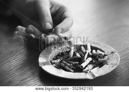 An Ashtray Filled With Cigarette Butts Stands On A Table, And A Hand Drops Ash From A Cigarette That