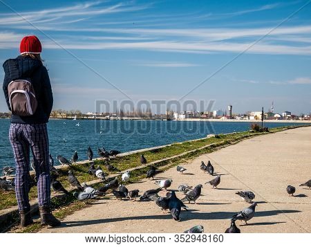 Girl With A Backpack Surrounded By Pigeons At The Edge Of A Lake, With Clear Blue Sky.