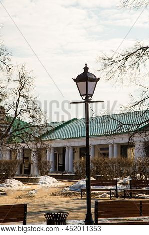 An Old Lantern Among The Benches Against The Background Of A Wall With Columns. Spring, Snow Melts, 