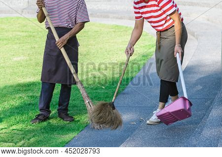 Female Worker Sweeping Yard With Broom Tool And Dustpan In Ornamental Backyard Garden. Woman Wear Ap