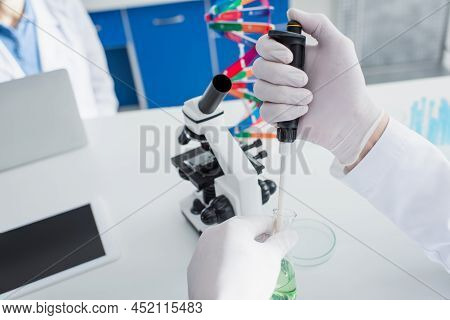Cropped View Of Biotechnologist In Latex Gloves Holding Micropipette And Flask Near Microscope.