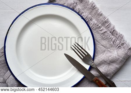 Old vintage white plate on table with fork and knife. Empty white plate. Emtpy plate on white background. Old rustic vintage plate with cutlery. Emtpy plate. Round white empty plate.