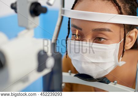 Female Patient In Medical Mask Getting Eyes Examined With Slit Lamp, Microscope That Allows Doctor T