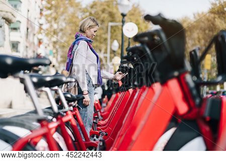 Blond Woman Taking A Red Bicycle In A Bike Rental Station In The City With Smartphone Sharing App - 