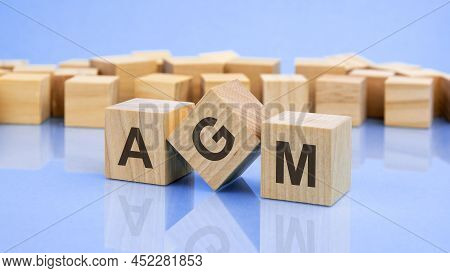 Three Wooden Cubes With The Letters Agm On The Bright Surface Of A Pale Lilac Table. The Inscription