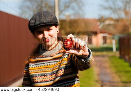 Defocus Ukrainian Young Smiling Woman In Hat Holding One Colored Red Egg On Nature Background. Easte