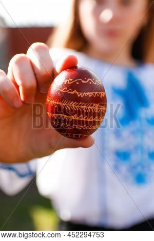 Defocus Ukrainian Girl In Vyshyvanka Holding One Colored Red Egg On Nature Background. Easter, Ukrai