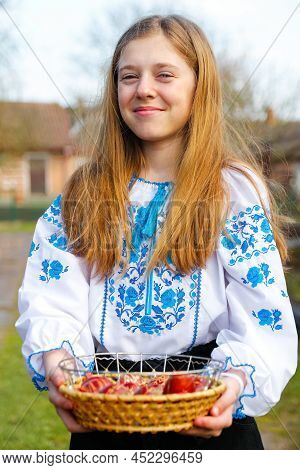 Defocus Ukrainian Girl In Vyshyvanka Holding Basket Of Colored Red Eggs On Nature Green Grass Backgr