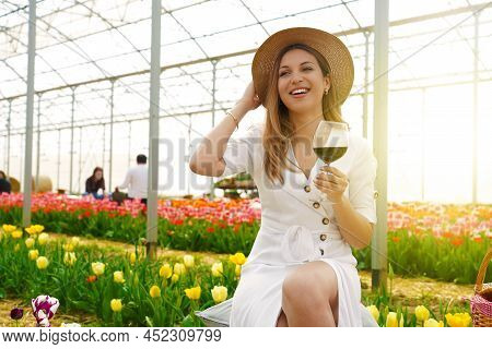 Relaxed Beauty Woman Holds Chalice Of Wine Between Tulips In Greenhouse
