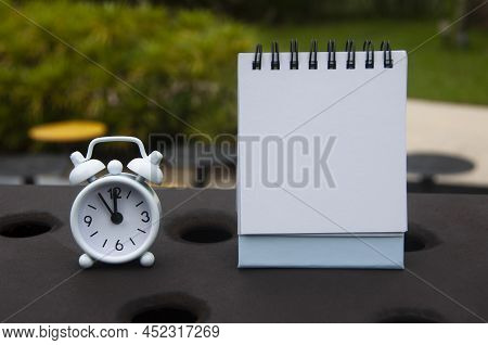 Alarm Clock Pointing At 11 O'clock With Blank Notepad. Blurred Park Background. Copy Space