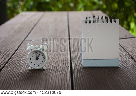 Alarm Clock Pointing At 1 O'clock With Blank Notepad On A Bench. Copy Space