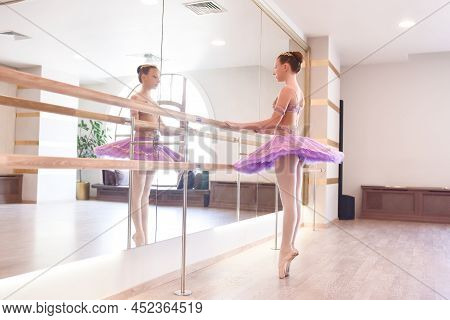 Young Ballerina Engaged In The Dance Studio. A Pretty Ballerina In A Purple Tutu Does Stretching Nea