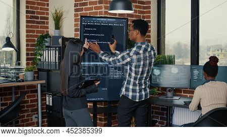 Software Engineers Analyzing Code On Wall Screen Tv Comparing Errors Using Digital Tablet Next To Pr