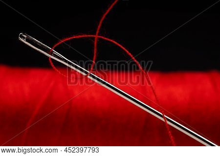 Eye Of The Needle With Red Polyester Thread. Spool Of Red Polyester Sewing Thread In The Background.
