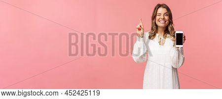 Portrait Of Lucky And Hopeful Beautiful Blond Girl In White Dress, Making Bet, Showing Mobile Phone 