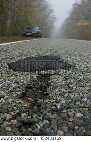 Potholes And A Patch On A Paved Road That Runs Through A Foggy Forest. There Is A Car Standing On Th
