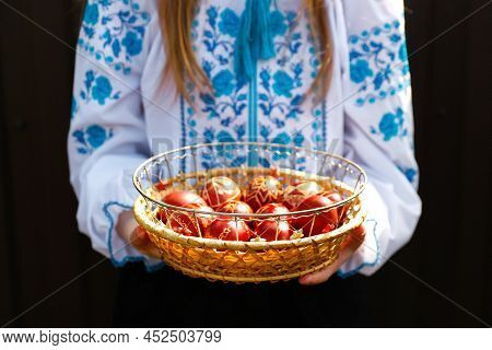 Defocus Ukrainian Girl In Vyshyvanka Holding Basket Of Colored Red Eggs On Black Background. Easter,