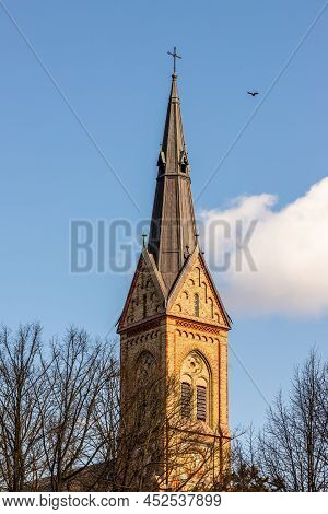 Dark Yellow Stone Church Tower, Leafless Spring Branches, Blue Sky With One Cloud And Black Bird. To