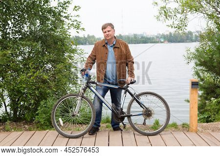 An Adult Blond Man In A Brown Corduroy Jacket Is Standing By The River In The Park With A Bicycle.