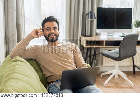 Front View Of Positive Hindi Man In Smart Casual Clothes Using Laptop While Sitting At The Sofa In H