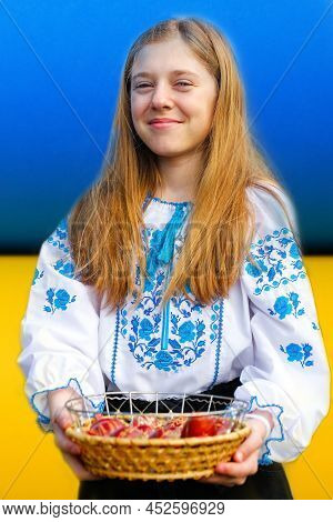 Defocus Ukrainian Girl In Vyshyvanka Holding Basket Of Colored Red Eggs On Ukrainian Flag Background