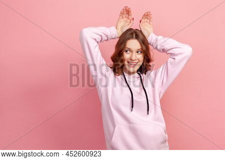 Portrait Of Carefree Happy Brunette Young Female In Hoodie Making Funny Bunny Ears With Hands On Hea