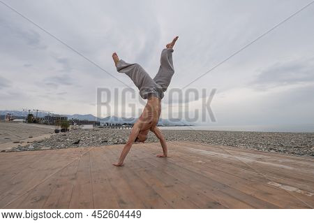 Shirtless Caucasian Man Doing Backflip On Pebble Beach.