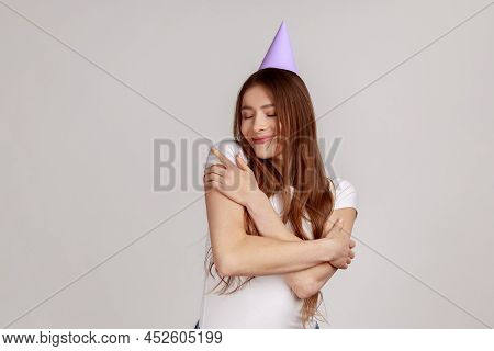 Attractive Dark Haired Woman In Party Cone On Head, Supporting, Congratulating And Embracing Herself