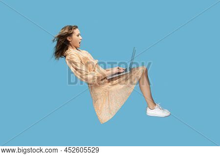 Hovering In Air. Surprised Girl In Yellow Dress Levitating, Looking At Laptop Screen Shocked Amazed,