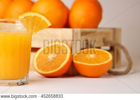 A Glass Of Fresh Orange Juice, A Slice Of An Orange On It, Wooden Box With Rope Handles Full Of Oran