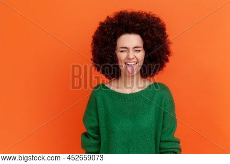 Positive Playful Woman With Afro Hairstyle Wearing Green Sweater Standing With Closed Eyes And Tongu