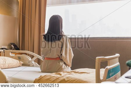 Back View Of Patient Woman Sitting On Bed In Hospital Ward Looking Out At Window