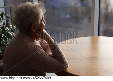 Elderly Woman Looking Out Of Window On Rainy Day, Space For Text. Loneliness Concept