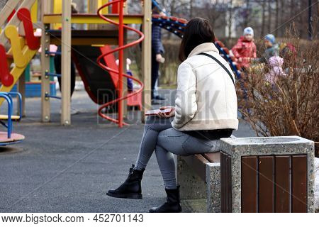 Girl In White Coat And Jeans Sitting With A Smartphone On The Bench On Playground In Spring Park. Co