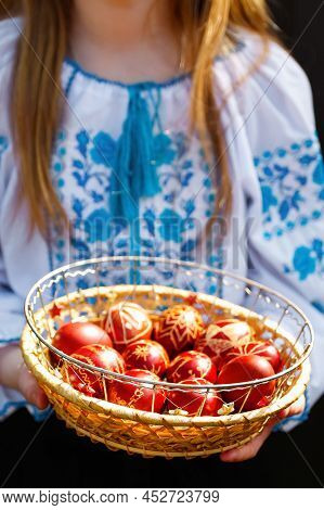 Defocus Ukrainian Girl In Vyshyvanka Holding Basket Of Colored Red Eggs On Nature Green Grass Backgr