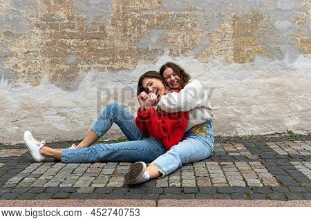 Two Best Friends Hugged Sitting On A Paved Sidewalk By An Old Wall