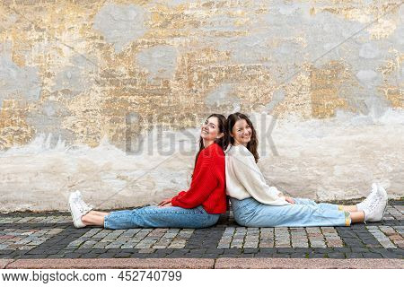 Two Young Women Sitting Back To Back On The Pavement By An Old Wall