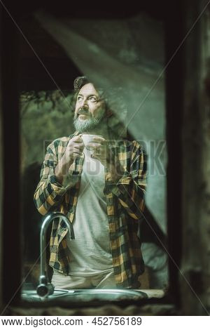 Mature Adult Man At Home Viewed From Outside Thrugh The Glass Winws Drinking Coffee And Looking Arou