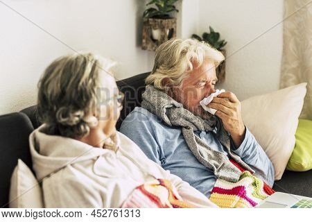Old Woman With Her Husband Sneezing Into Tissue Paper At Home. Senior Couple Relaxing On Sofa In Sic