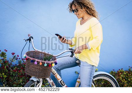Colorful Yellow And Blue Image Of Young Woman Using Cell Phone Sitting On A Cute Bike And Blue Wall 